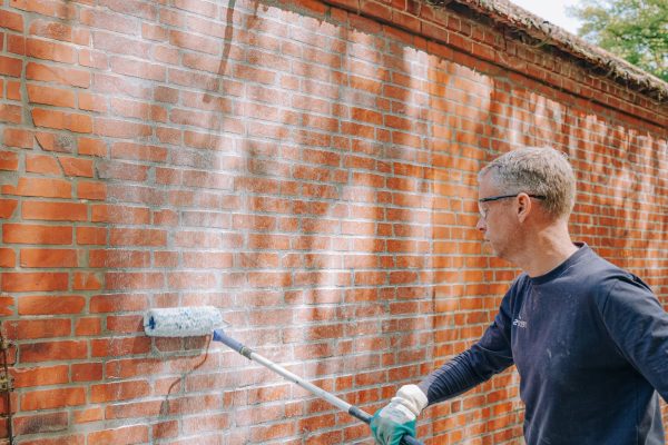 Handwerker trägt PFAS-frei formulierte Funcosil Creme mit einer Farbrolle auf eine rote Backsteinwand auf.