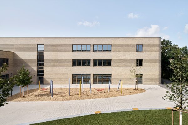 Moderne Außenansicht der Schule mit heller Klinkerfassade und einem Spielplatz mit Hängematten im Vordergrund.