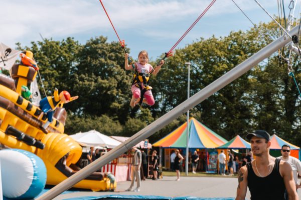 Kind springt hoch hinaus im Bungee-Seil auf einem bunten Jahrmarkt mit Zelten und einer großen Hüpfburg.