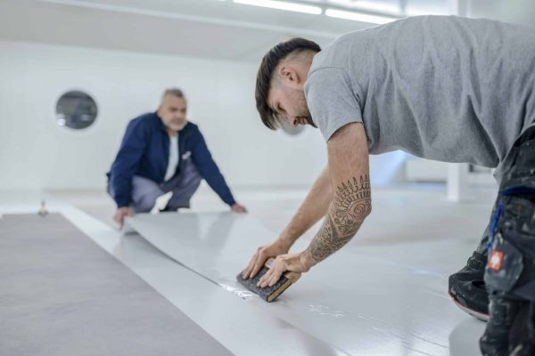 Zwei Handwerker bei der fachgerechten Verlegung UZIN Plurafilm Hydro auf einem hellen Hallenboden.