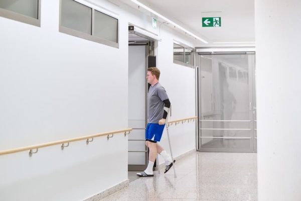 Mann auf Krücken nutzt barrierefreie Türen in Flucht- und Rettungswegen im Krankenhaus mit Notausgangsschild.