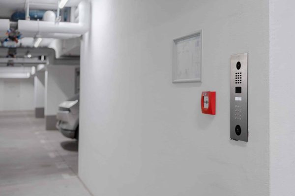 Moderne Türsprechanlage aus Edelstahl an einer weißen Wand in der Garage neben einem Feuermelder.