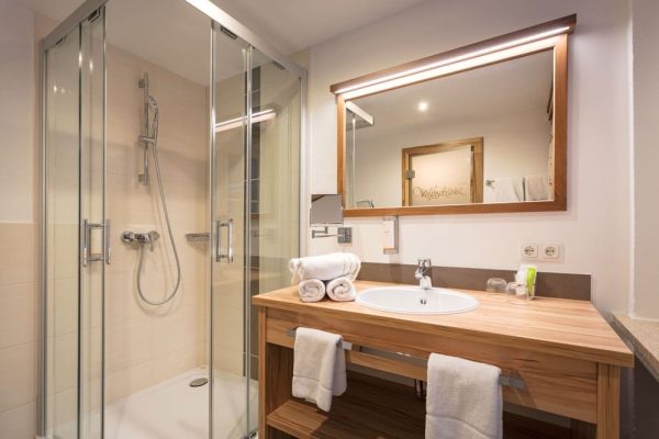 Modernes Bad im Sport- und Wellnesshotel Waldschlößl mit eleganter Glasdusche und rustikalem Holzwaschtisch.