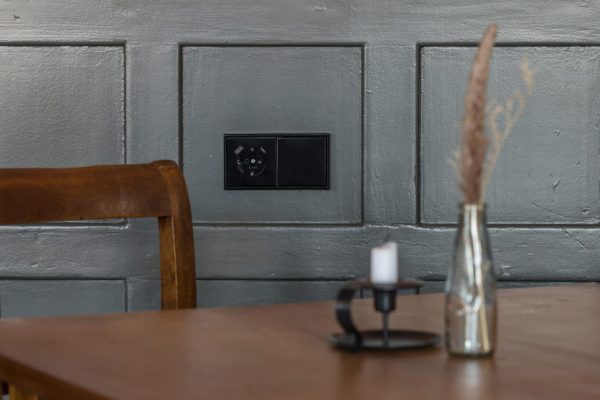 JUNG Schalter und USB-Steckdose LS 990 in Graphitschwarz matt an grauer Wand über einem Holztisch.