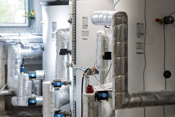 Drei weiße Optima Energiespeicher in einer Heizzentrale mit isolierten Rohrleitungen für moderne Wärmepumpen.