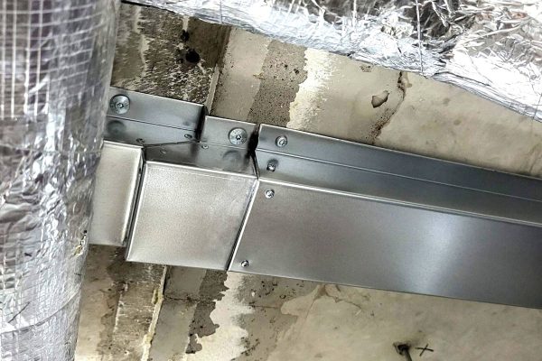 Metallischer Lüftungskanal an einer Betondecke mit einer kleinen Stufe am Übergang der Bauteile.