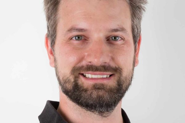 Mark Iten lächelt freundlich mit Bart und schwarzem Poloshirt vor einem neutralen weißen Hintergrund.