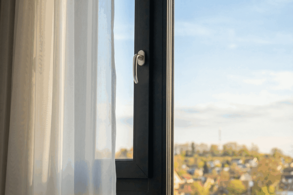Anthrazitfarbenes REHAU ARTEVO Fenster mit weißem Vorhang und Blick auf eine sonnige Hügellandschaft.