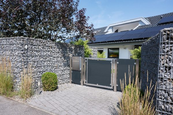 Moderner Zaunbriefkasten aus Edelstahl mit Trapez-Dach an einer Gabionenwand vor einem Haus mit Solaranlage.
