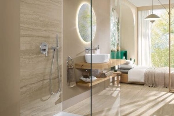 Ecora Hotelbad mit offener Glasdusche, rundem Leuchtspiegel und Holzwaschtisch in warmem Naturstein-Look.