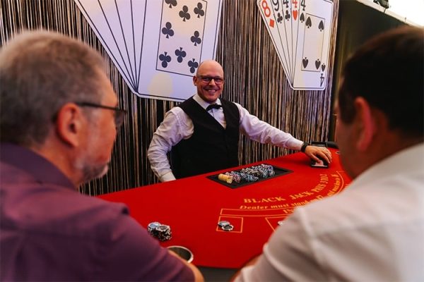 Eine Runde Black Jack gefällig? Ein lächelnder Dealer am roten Spieltisch mit Jetons und zwei Spielern.