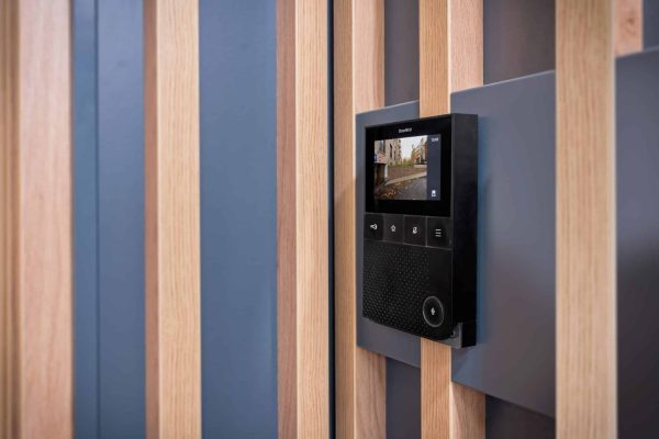 Moderne DoorBird Innenstation an einer Wand mit Holzlamellen zeigt das Kamerabild der Haustür.