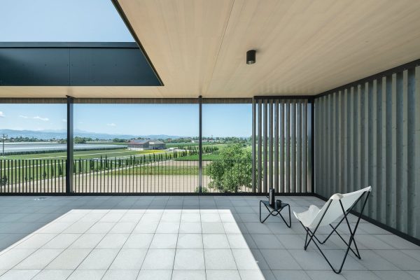 Moderner Balkon mit Decke aus CLT, weißem Klappstuhl und weitem Ausblick auf grüne Felder.