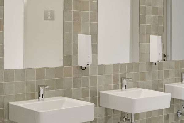 Moderne weiße Waschbecken und Spiegel im Sensoria Museum Holzminden vor hochwertigen grünlich-grauen Fliesen.