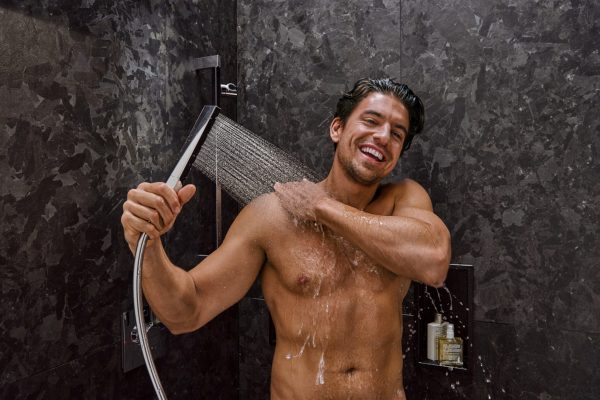Lächelnder Mann genießt das Wasser einer Handbrause vom hansgrohe Duschexperte vor dunklen Natursteinfliesen.