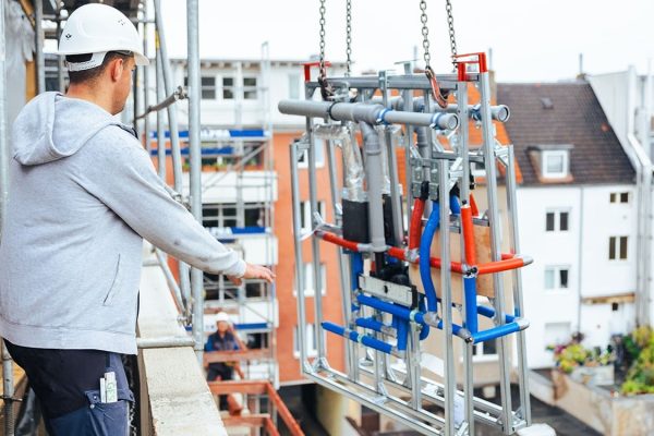 Anlieferung einer Installationswand TECEsystem per Kran am Rohbau mit Bauarbeitern auf einem Gerüst.