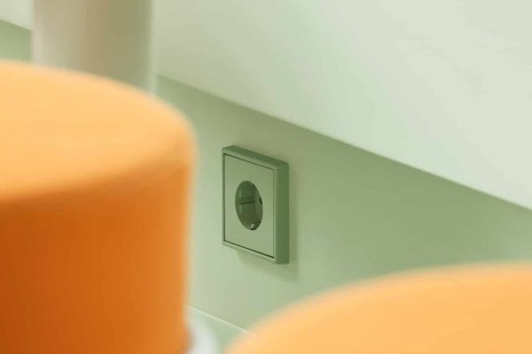 JUNG SCHUKO Steckdose LS 990 in vert anglais pâle an grüner Wand hinter orangefarbenen Hockern.