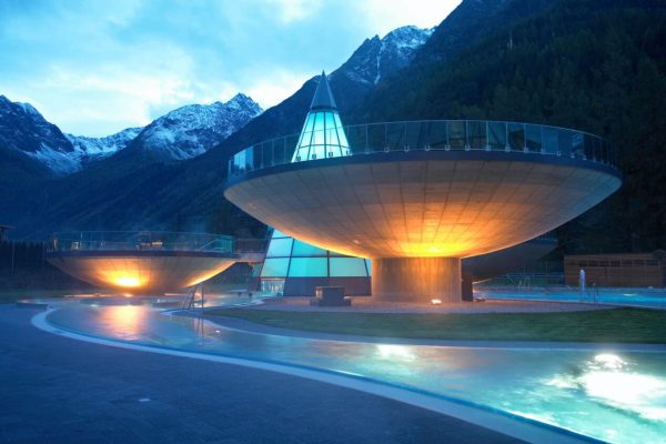 Aqua Dome Tirol Therme Längenfeld mit beleuchteten Schalenbecken bei Abendstimmung vor einem Bergpanorama.