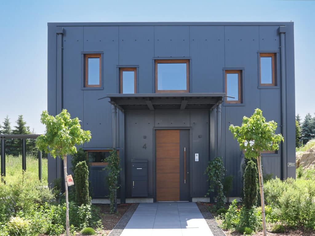 Moderne Hausfassade eines Green Steel Home mit dunkelgrauen Stahlpaneelen, Holzrahmenfenstern und begrüntem Vorgarten unter blauem Himmel.