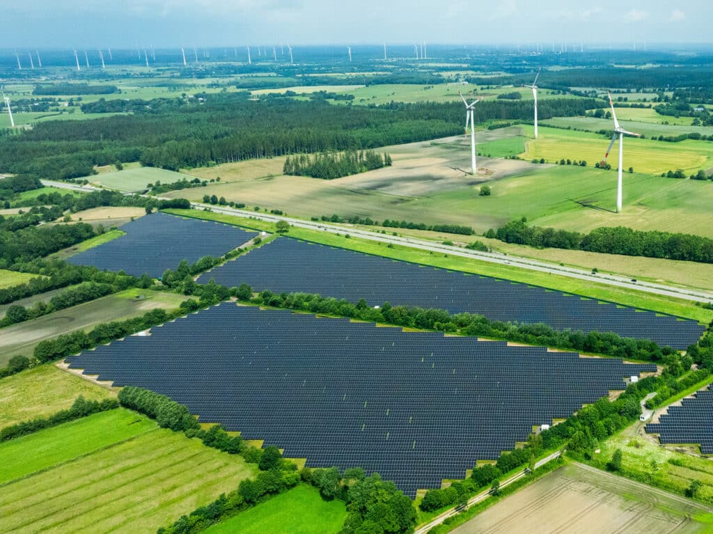 Luftaufnahme eines großen Solarparks inmitten grüner Landschaft mit angrenzenden Windkraftanlagen.