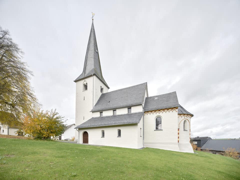 Außenansicht einer kleinen Kirche mit spitzem Turm, hellem Putz und umgebender Grünfläche