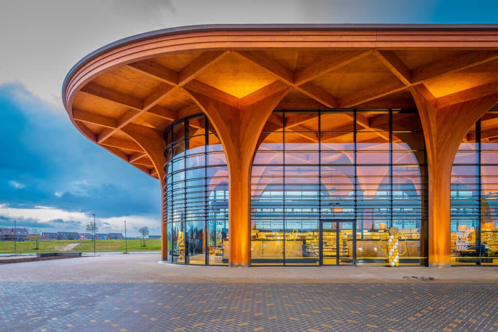 Moderne Supermarktfassade mit markanter Holzkonstruktion, großen Glasflächen und geschwungenem Dach, beleuchtet in warmem Licht.