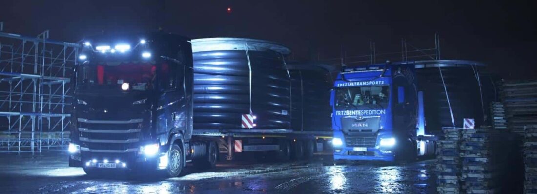 Zwei Schwerlast-Lkw transportieren bei Nacht große Kabeltrommeln mit vorisolierten Fernwärmerohren. Die Scheinwerfer leuchten die nasse Fahrbahn.