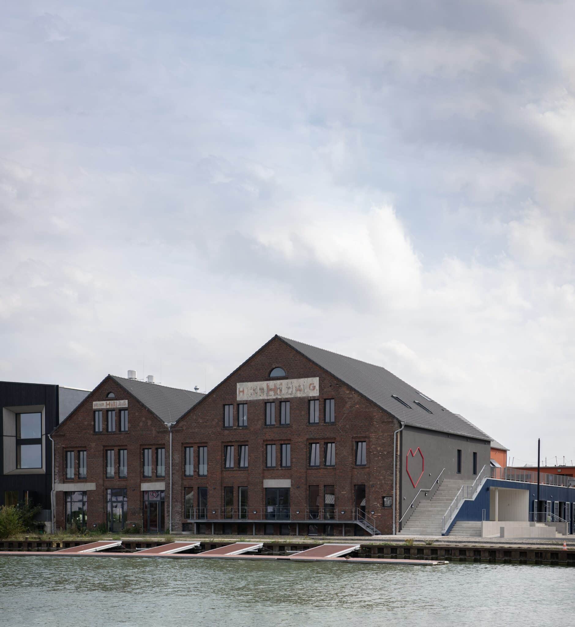 Außenansicht des historischen Hill-Speichers in Münster, modern saniert mit roter Backsteinfassade, großen Fenstern und markantem Herzsymbol an der Seitenwand, direkt am Wasser gelegen.