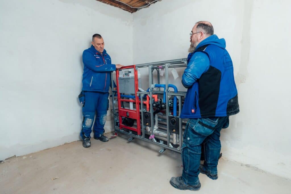Zwei Installateure positionieren ein TECE-Vorwandelement mit Leitungen und Anschlüssen in einem unfertigen Badezimmer während der Renovierungsarbeiten.