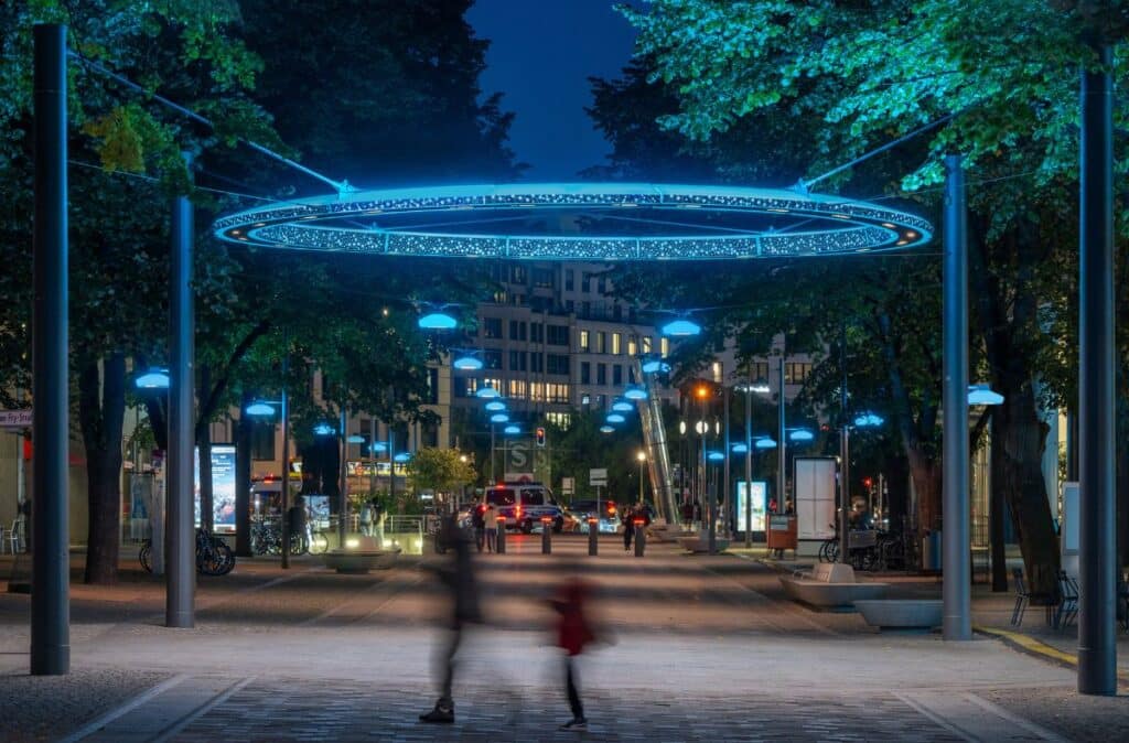 Abendszene am Potsdamer Platz in Berlin mit leuchtend blauen Ringleuchten und Passanten in Bewegung.