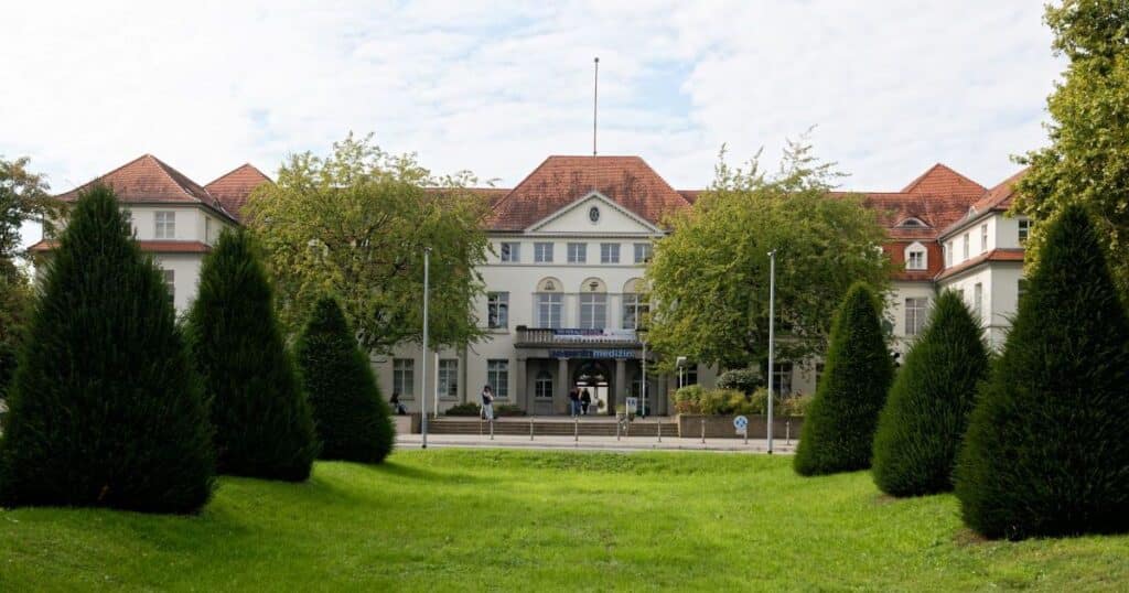 Frontansicht des Hauptgebäudes der Universitätsmedizin Mainz mit rotem Dach, begrüntem Vorplatz und symmetrisch angeordneten Bäumen.