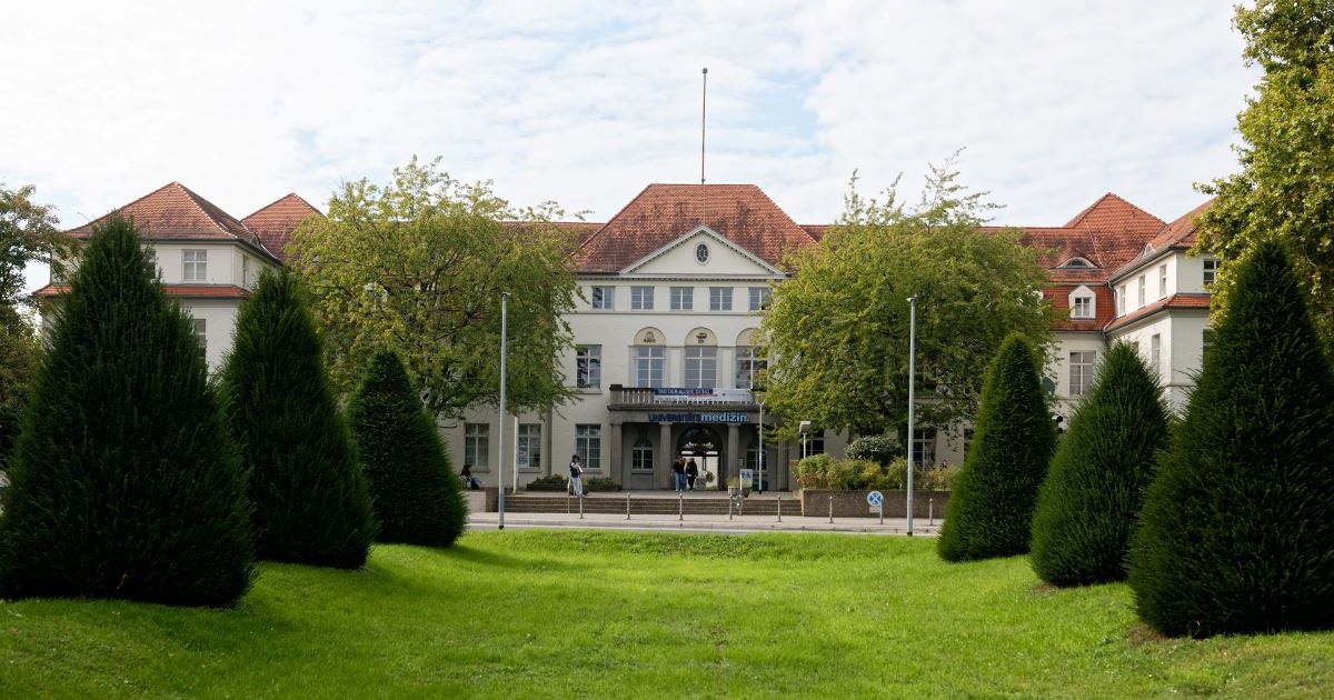 Frontansicht des Hauptgebäudes der Universitätsmedizin Mainz mit rotem Dach, begrüntem Vorplatz und symmetrisch angeordneten Bäumen.