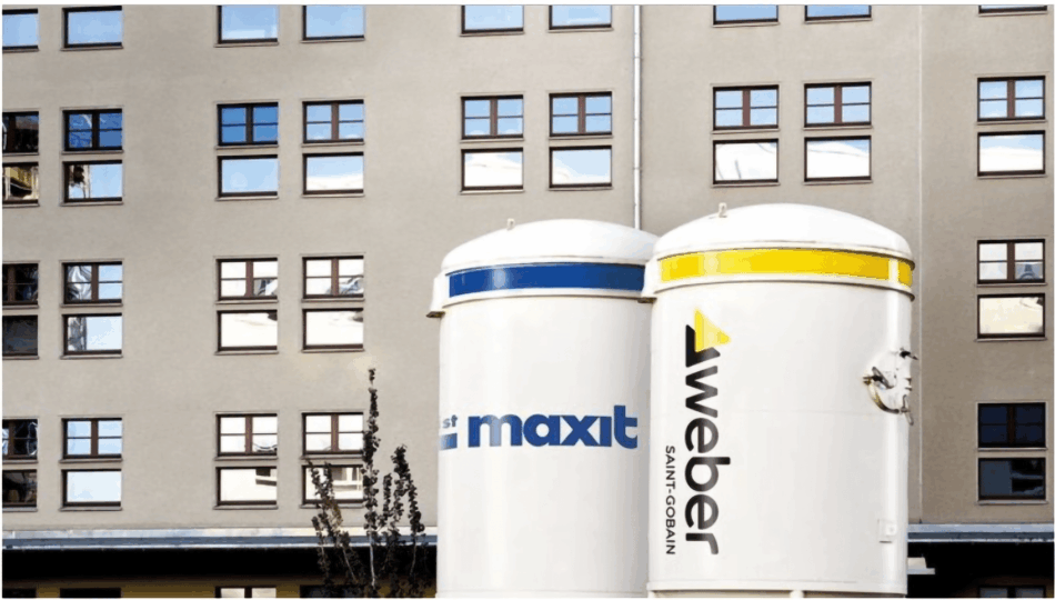 Zwei Silos mit den Logos von maxit und Saint-Gobain Weber vor einem Wohngebäude.