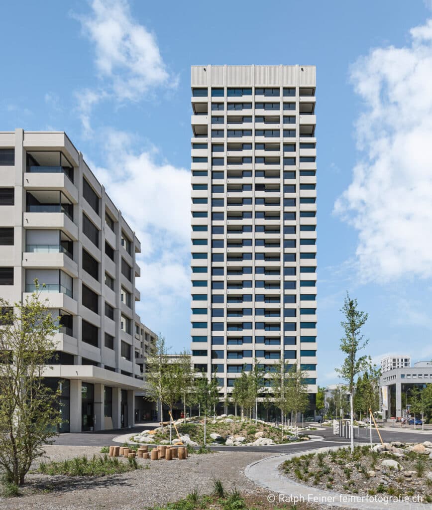 Modernes Wohnhochhaus in Zürich mit klar gegliederter Betonfassade, begrüntem Vorplatz und blauem Himmel im Hintergrund